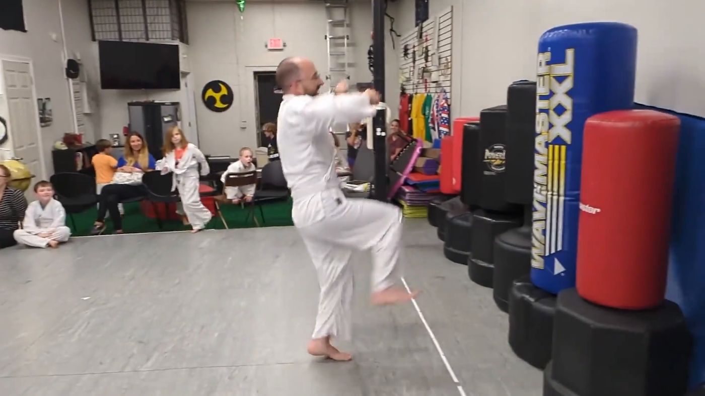A student lands a flying kick on a heavy bag while classmates watch